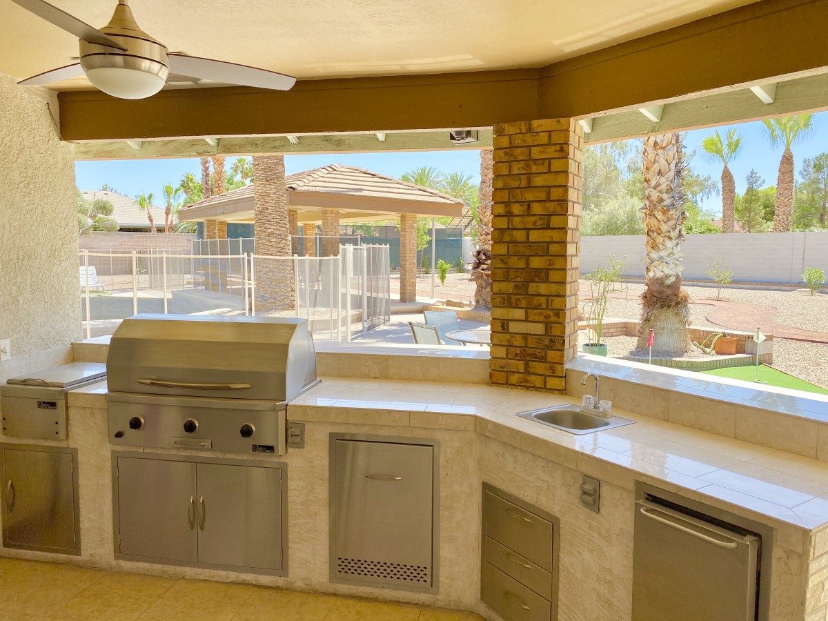 A spacious outdoor kitchen with a barbecue grill and brick pillars overlooking a sunny backyard with palm trees.