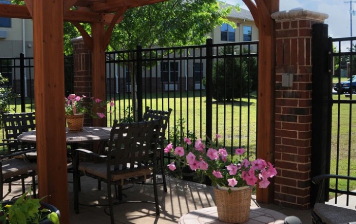 A cozy patio with a pergola, adorned with flowers and outdoor furniture, overlooking a fenced garden.