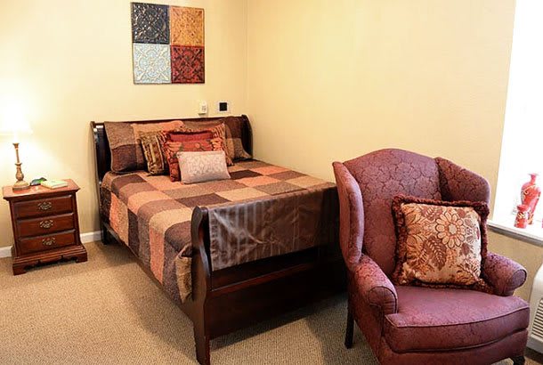 A cozy bedroom with a quilt-covered bed and an armchair, featuring decorative pillows and wall art.