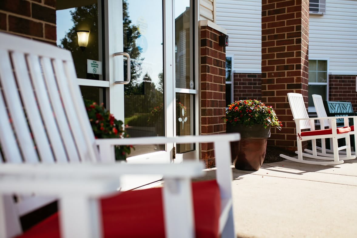 A sunny patio area with white chairs and potted plants outside a brick building.