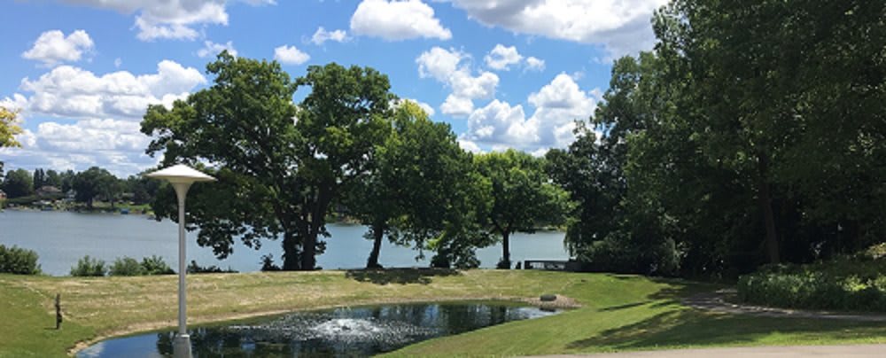 A serene lakeside scene with lush trees, a small fountain, and a partly cloudy sky.