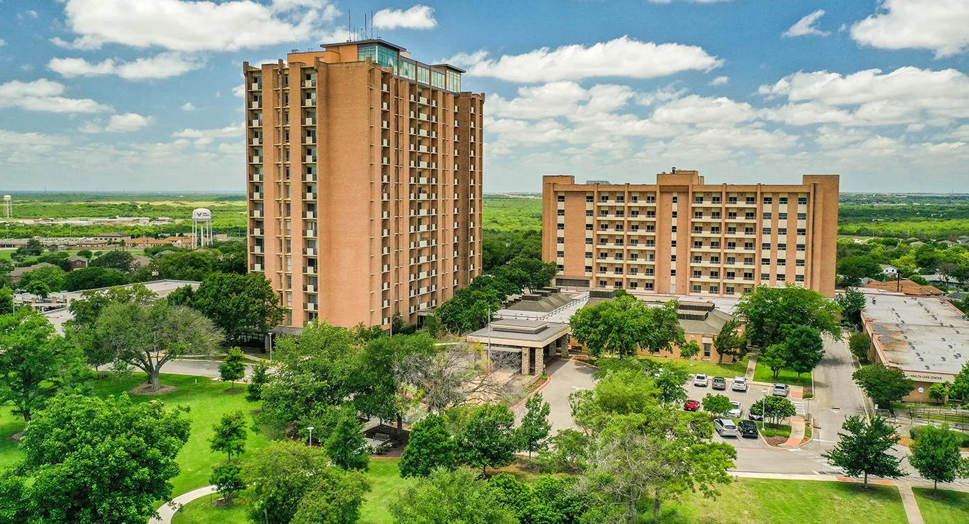 A view of two high-rise residential buildings surrounded by greenery and trees.