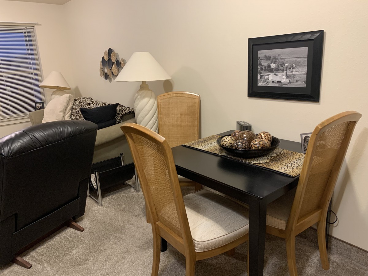 A cozy living room with a small dining area, featuring wooden chairs, a table, lamps, and wall artwork.