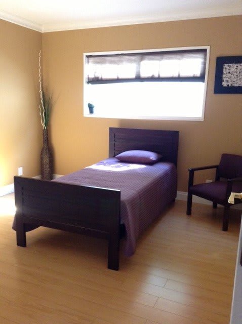 A minimalist bedroom with a single bed featuring purple bedding, wooden furniture, and natural lighting.