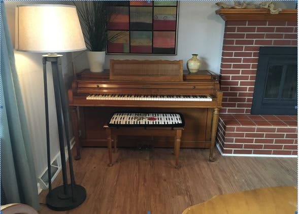 A cozy living room corner featuring a piano with a bench, a nearby floor lamp, and a brick fireplace.
