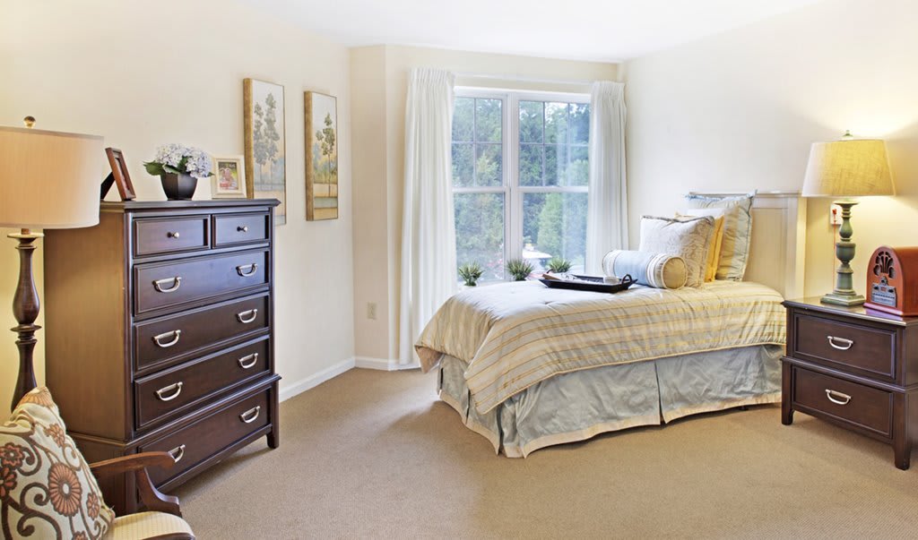 A cozy bedroom with a neatly made bed, dresser, and a large window allowing natural light to fill the space.