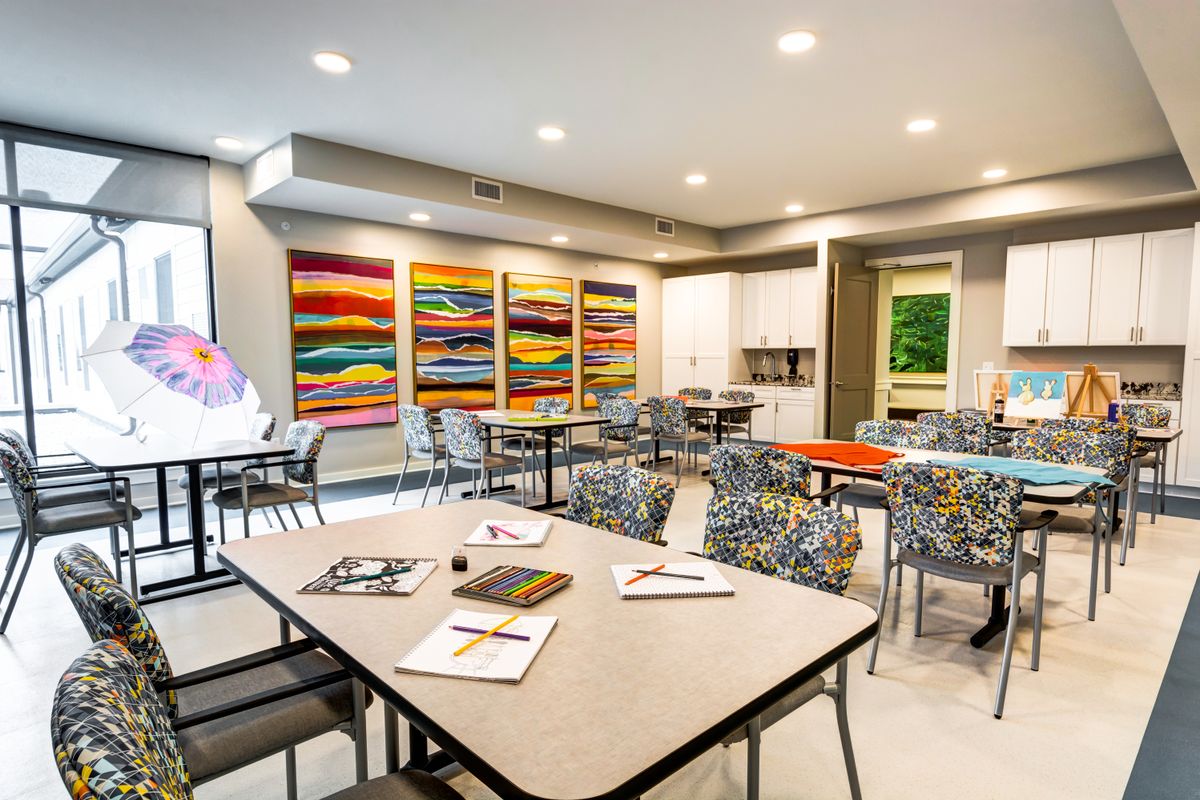 A brightly lit art studio with colorful paintings on the walls, tables with art supplies, and patterned chairs.