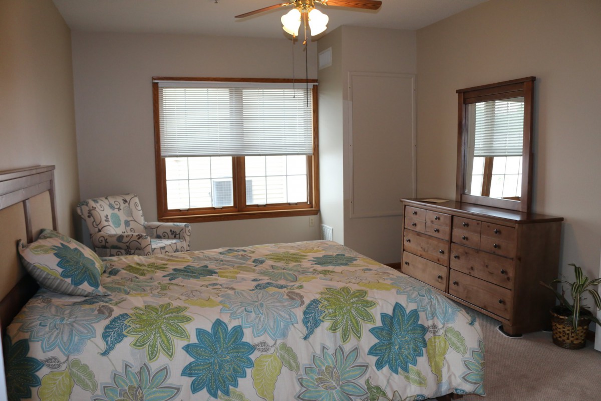 A cozy bedroom with a floral bedspread, wooden dresser, and large windows with blinds.