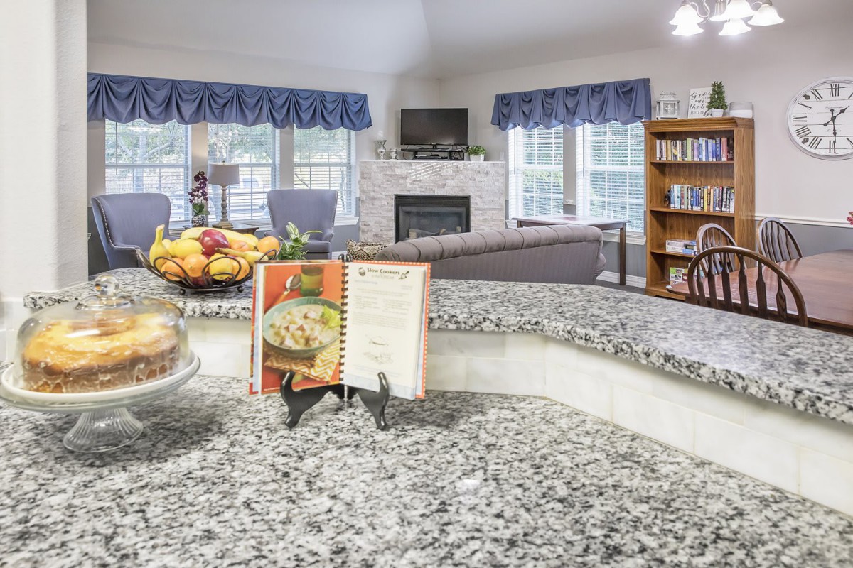 A cozy kitchen with a cookbook on the countertop, overlooking a living room with a fireplace and bookshelf.