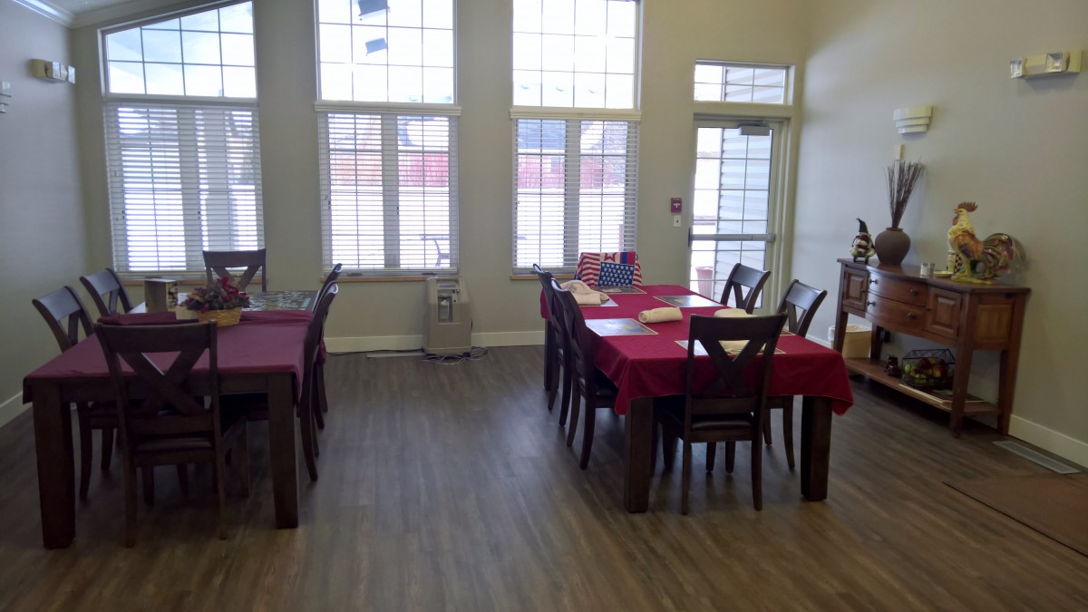A bright dining room with tables covered in red tablecloths, surrounded by wooden chairs and illuminated by large windows.