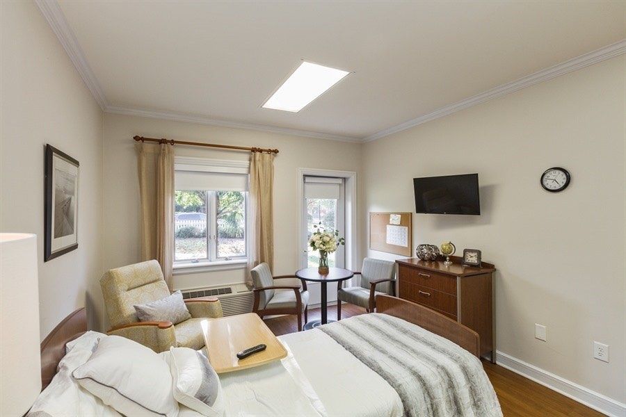 A cozy bedroom with a bed, chairs, and a television, featuring modern decor and natural light from the windows.
