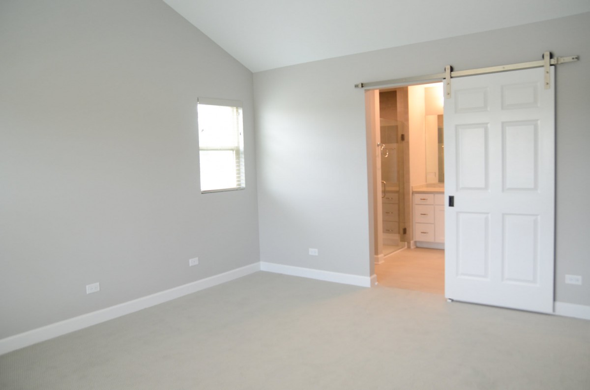 An empty room with gray walls, carpet flooring, a window, and a doorway leading to a bathroom.