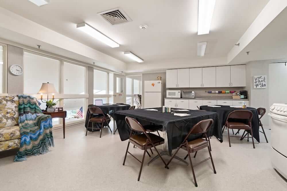 A well-lit community kitchen with tables and chairs arranged, featuring large windows and a row of cabinets.
