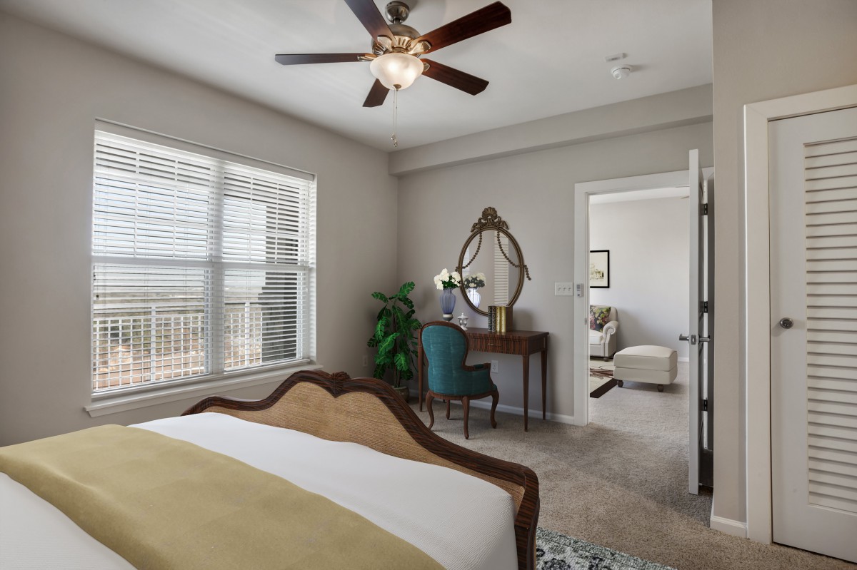 A beautifully furnished bedroom featuring a comfortable bed, vintage-style desk with a mirror, and a ceiling fan. The room is brightened by natural light streaming through the large window.