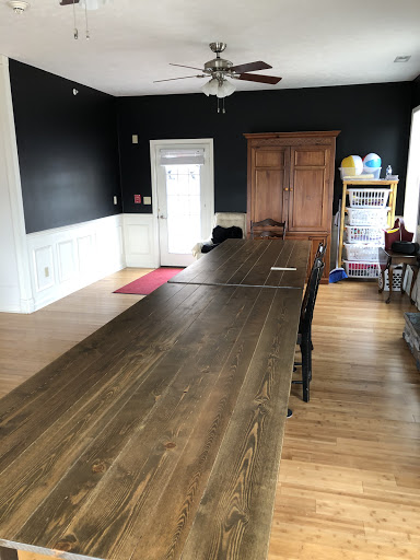 A spacious interior room featuring a long wooden table, black walls, and a warm wooden floor.