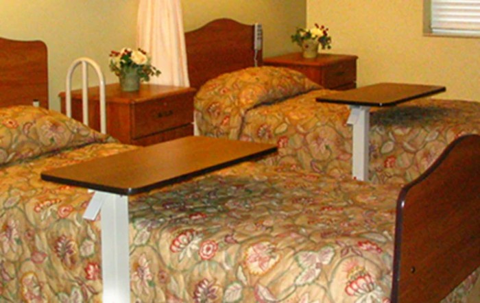 A hospital room featuring two beds with overbed tables and flower arrangements.