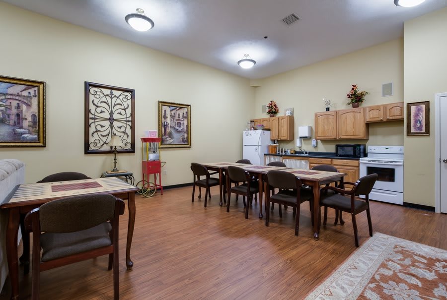 A spacious kitchen and dining area with wooden flooring, featuring multiple tables and chairs, a refrigerator, microwave, and decorative artwork on the walls.