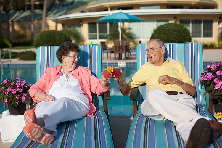 An elderly couple relaxes by the pool, sharing a toast with refreshing drinks.
