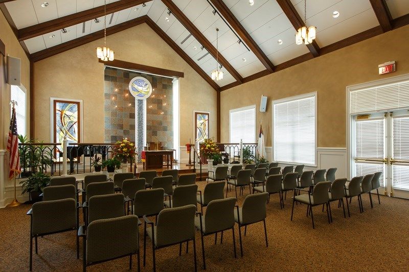 An interior view of a chapel with rows of seating facing an altar, featuring large windows and decorative lighting.