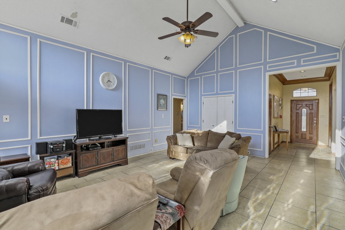 A cozy living room with blue paneled walls, featuring a sofa set, television, ceiling fan, and wooden flooring.