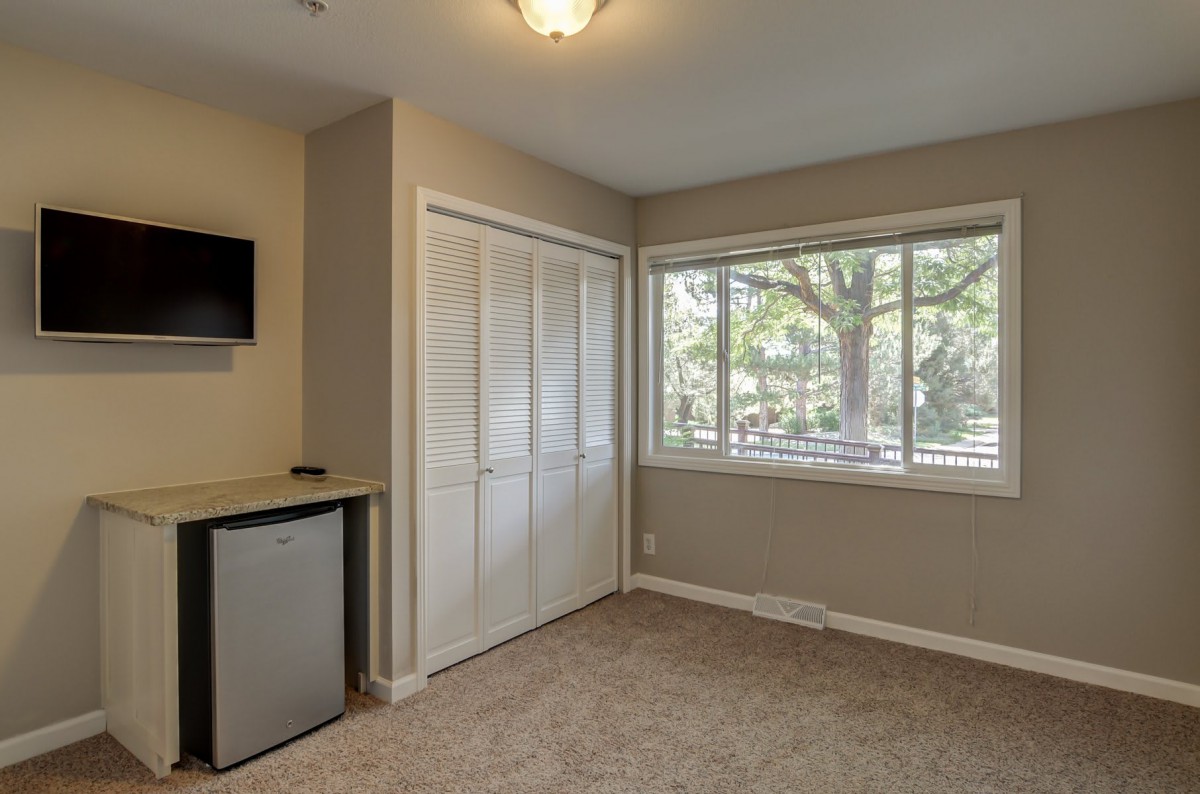 A cozy room with a large window, carpet flooring, a closet, wall-mounted TV, and a mini fridge.
