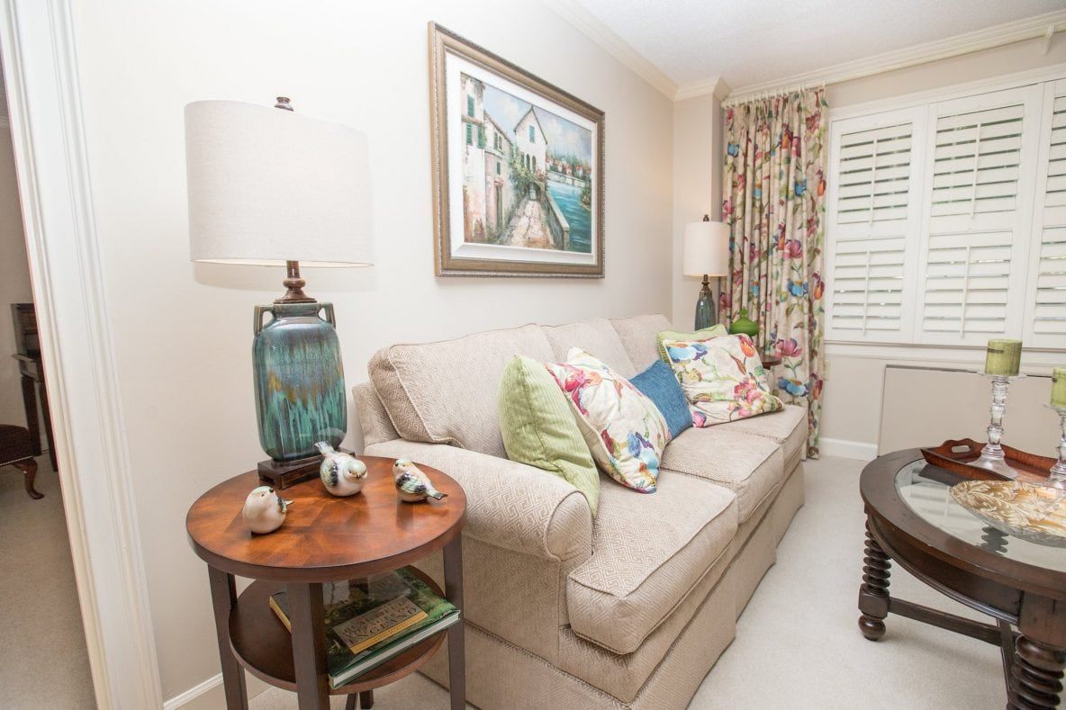 A cozy living room with a beige sofa adorned with colorful cushions, a side table with a lamp, and decorative elements.