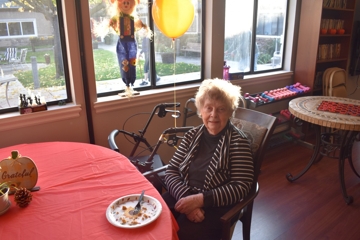An elderly woman sitting at a table with a balloon nearby, enjoying a birthday celebration indoors.