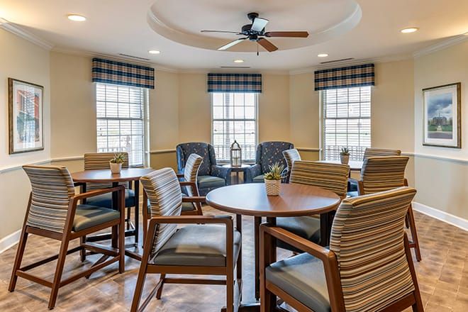 A cozy seating area with tables and chairs, featuring large windows and a ceiling fan.