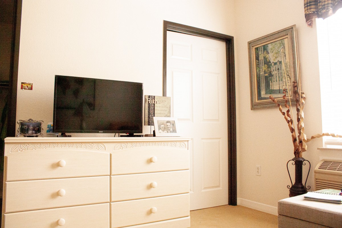 A cozy bedroom corner featuring a white dresser with a TV on top, adjacent to a door and a framed mirror on the wall.