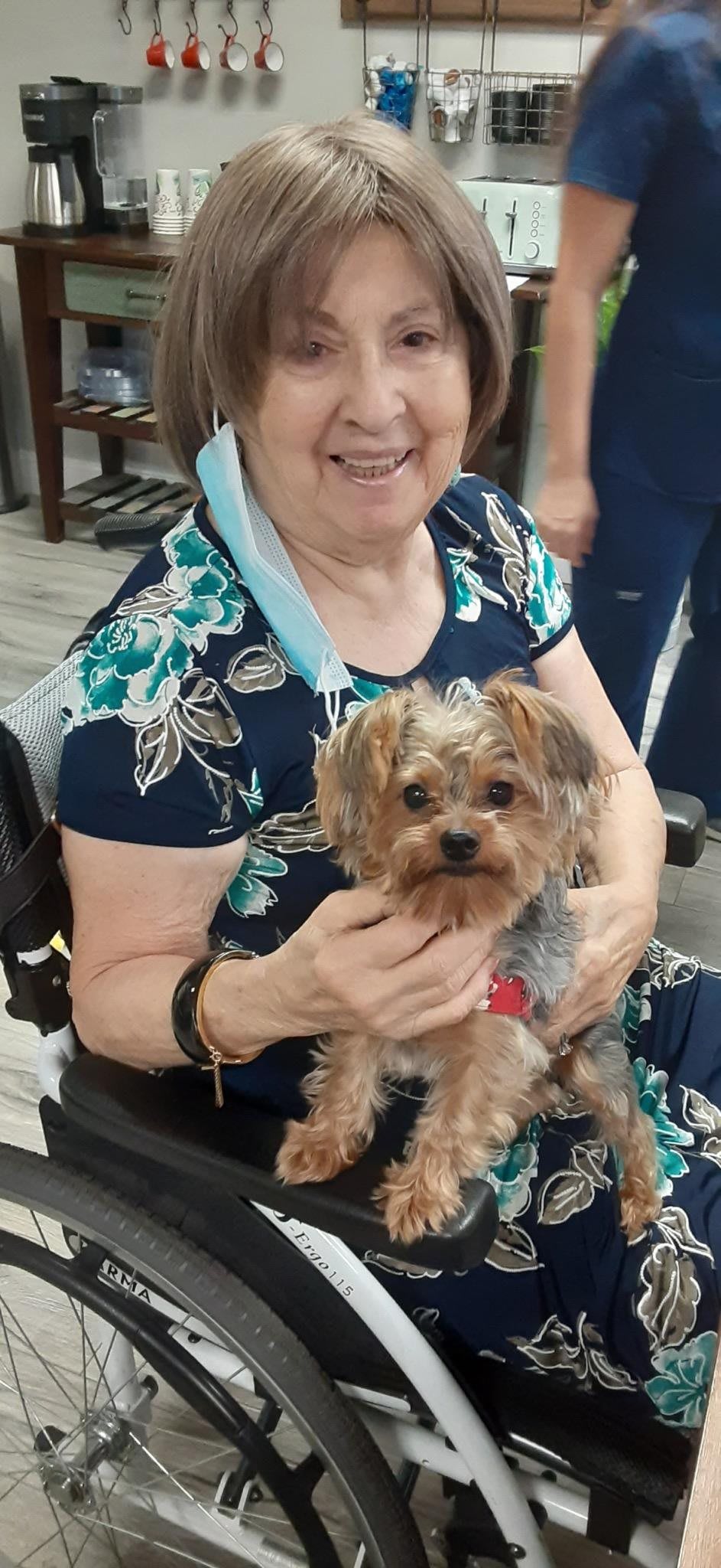 A woman sitting indoors, smiling while holding a small dog on her lap.