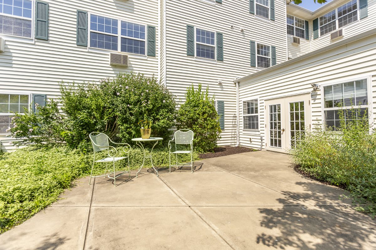 A sunny patio area with a small table and chairs, surrounded by lush greenery next to a residential building.