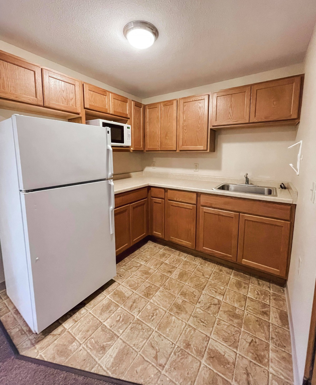 A compact apartment kitchen with wooden cabinets, a refrigerator, microwave, and tiled flooring.