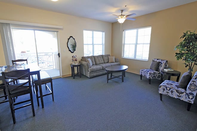 A cozy living room with a sofa, chairs, and tables under natural light from large windows.
