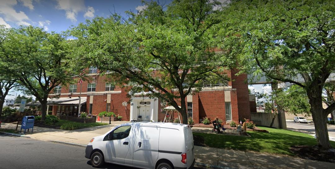 A street view of a brick building with a large tree in front, a white van parked nearby, and people sitting on benches.