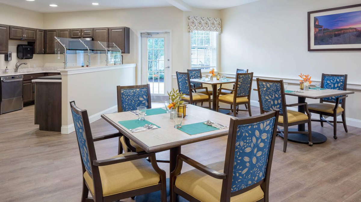 A well-lit dining area with neatly arranged tables and chairs, featuring a view into the kitchen.