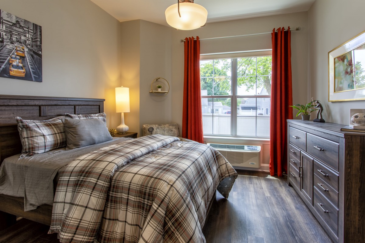 A cozy bedroom with plaid bedding, a large window with orange curtains, and warm lighting.