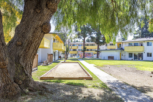 A serene view of an apartment complex featuring yellow buildings, a bocce ball court, and lush green landscaping.