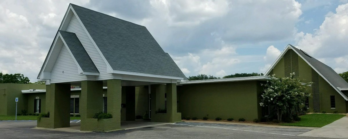 A green building with a sloped roof and a parking area under a cloudy sky.
