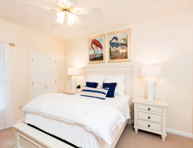 A bright bedroom with white furnishings, featuring a cozy bed, nightstands with lamps, and colorful artwork on the wall.