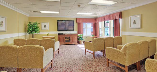A well-furnished living room with multiple chairs facing a television, featuring large windows and patterned carpet.