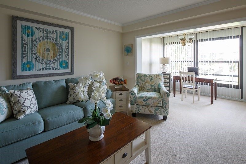 A cozy living room with a blue sofa, patterned armchair, and wooden coffee table. The room is decorated with artwork, cushions, and includes a view of the adjacent dining area.