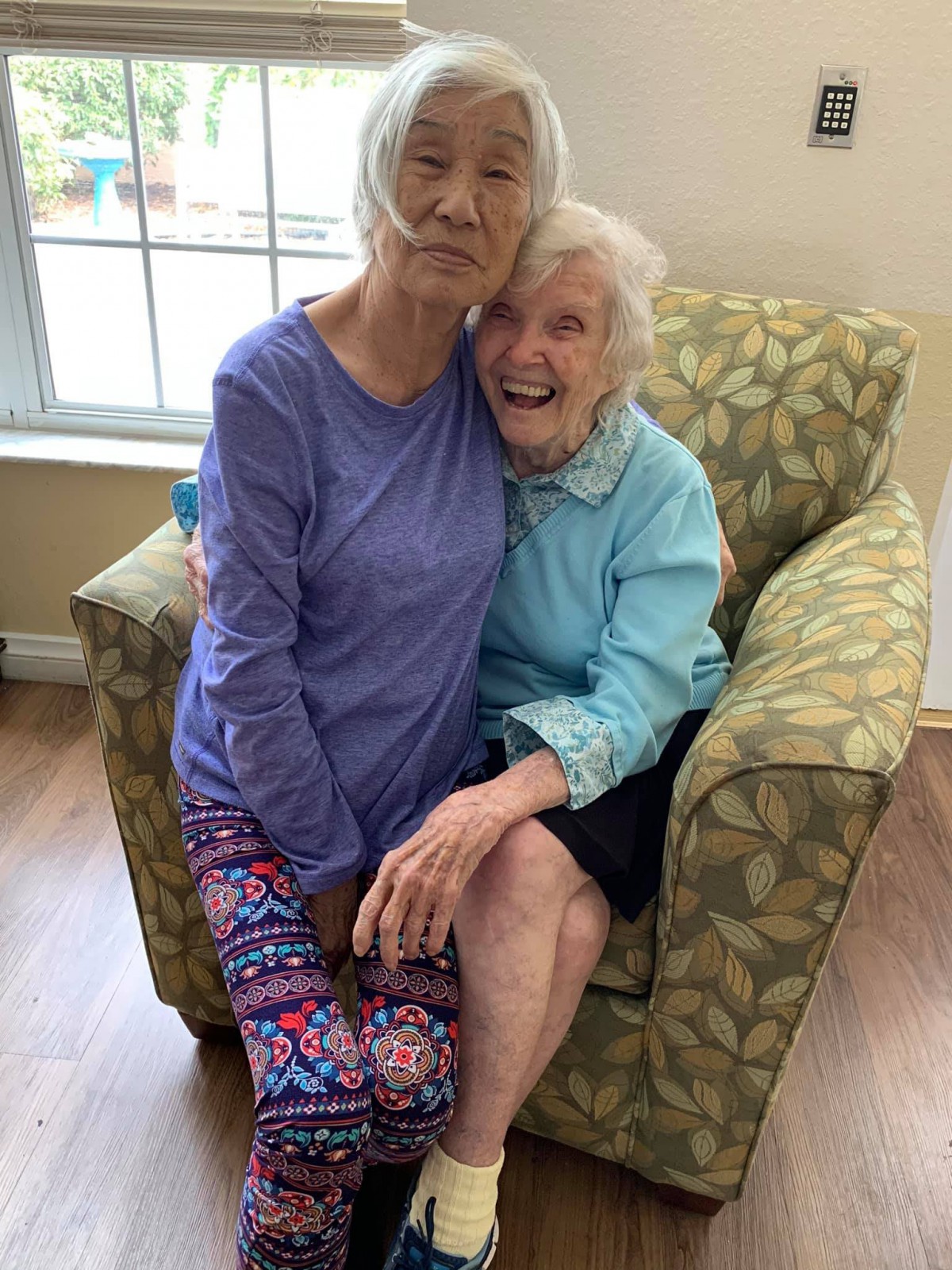 Two elderly women share a joyful embrace while seated indoors.
