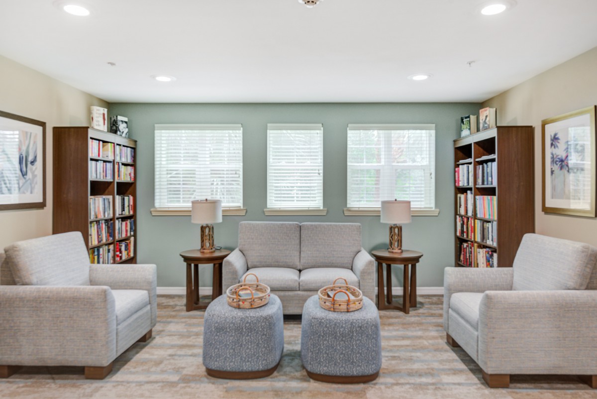 A cozy living room with bookshelves, a sofa, armchairs, and ottomans under a set of three windows.