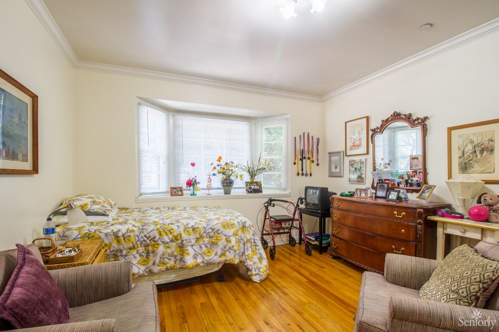 A cozy bedroom with a large window, decorated with plants and vintage furniture on a hardwood floor.