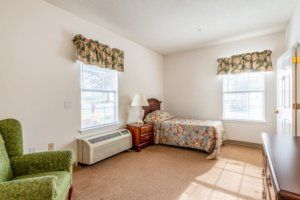 A cozy bedroom with a single bed, two windows with floral curtains, and a bedside table with a lamp.