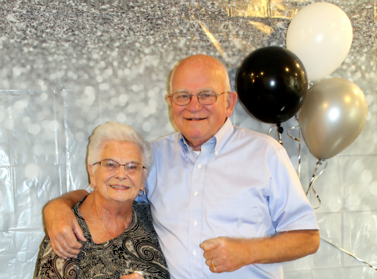 An elderly couple smiling together in front of a festive backdrop with balloons.