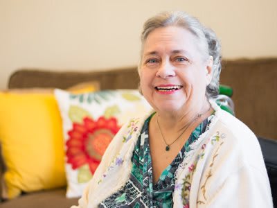 An elderly woman smiling while sitting indoors with colorful pillows in the background.