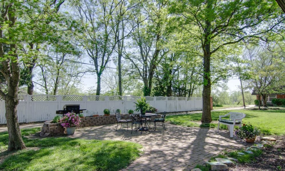 A sunny backyard patio with outdoor seating surrounded by trees and a white fence.