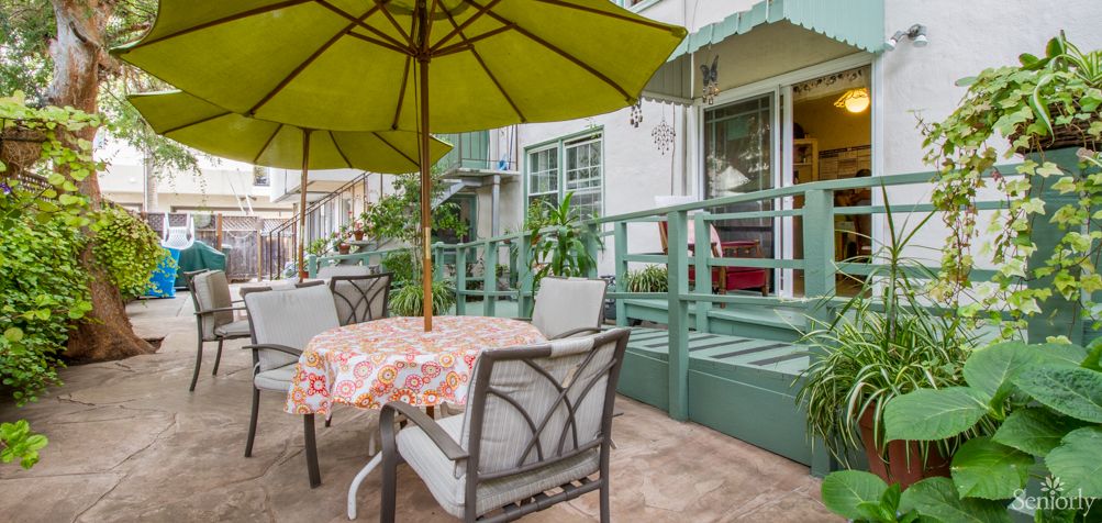 A cozy outdoor patio setup with a table and chairs under a green umbrella, surrounded by greenery.
