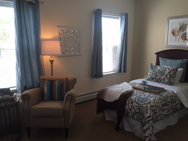 A cozy bedroom with a neatly made bed, soft lighting from an elegant lamp, and comfortable seating next to a window.